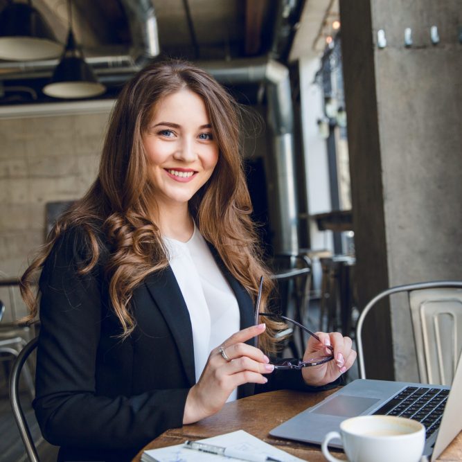 Widely smiling businesswoman working on laptop sitting in a cafe. Woman with long hair wears black jacket and white blouse and holds black eyeglasses in hands.